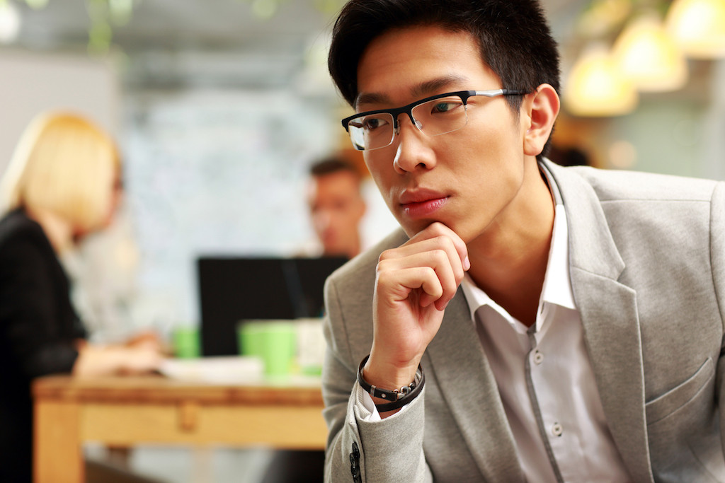 Portrait of a pensive asian man sitting in office, with his colleagues in background